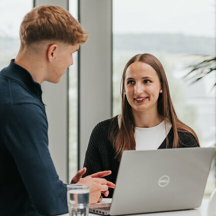 Zwei junge Erwachsene sprechen miteinander. Vor ihnen steht ein aufgeklappter Laptop.
