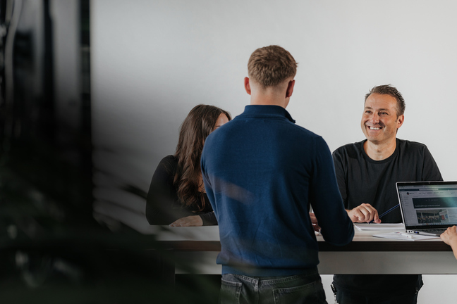 3 Personen, zwei Männer und eine Fau an einem Stehtisch, freundlich lächelnd