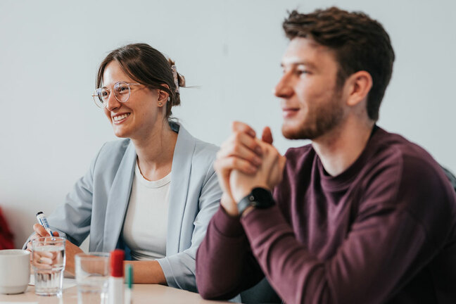 Zwei Personen sitzen an einem Tisch. Die Frau ist im Fokus, der Mann ist unscharf.