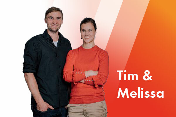 Zwei Personen stehen nebeneinander und schauen freundlich in die Kamera. Ein Mann hat ein schwarzes Hemd an. Eine Frau steht rechts daneben und trägt eine rote Bluse.