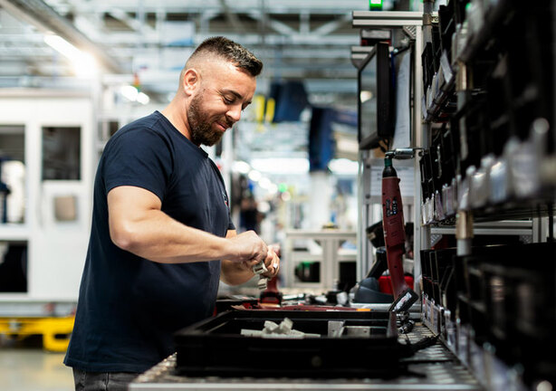 Ein Mann arbeitet an einer Werkbank in einer industriellen Umgebung. Sie hantiert mit Werkzeugen und Ausrüstung. Im Hintergrund sind Maschinen und andere industrielle Elemente zu sehen.