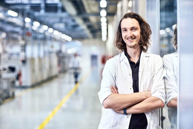 Ein Mann steht angelehnt an einer Wand und schaut lachend in die Kamera. Er trägt einen weißen ESD-Kittel und im Hintergrund sieht man eine Produktionslinie.