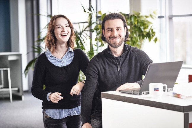 Zwei Personen stehen in einem modernen Büroumfeld und schauen lachend in die Kamera.