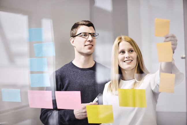 Zwei Personen stehen vor einer Glaswand mit vielen bunten Haftnotizen.