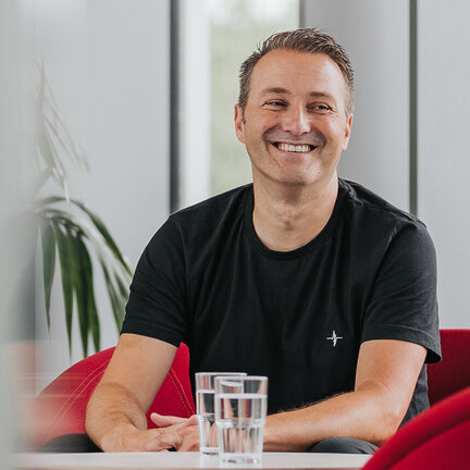 Ein Mann mit einem schwarzen Shirt sitzt lachend auf Lounge-Stühlen.