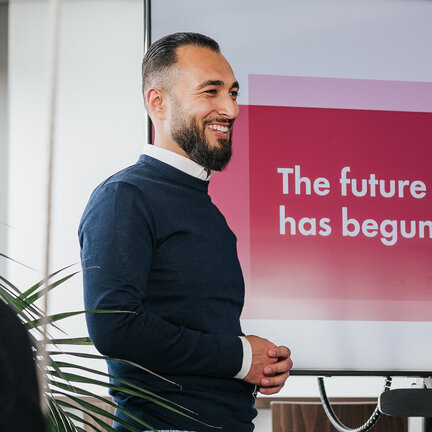 Ein Mann steht vor einem Bildschirm mit der Aufschrift "The future has begun". Er trägt einen dunkelblauen Pullover und hellblaue Jeans.
