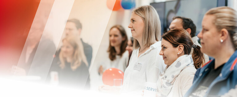 Eine Gruppe von Menschen in einer sozialen oder beruflichen Umgebung. Einige halten rote Luftballons. Der Hintergrund ist innen und mit Dekorationen, einschließlich Luftballons, versehen.